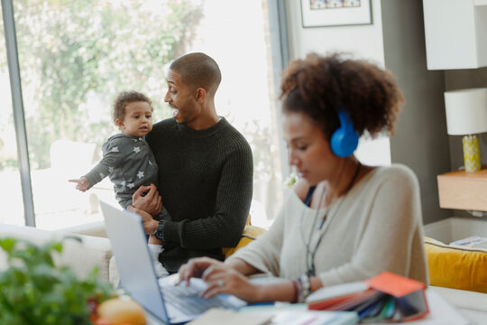Father holding baby daughter behind working mother at laptop