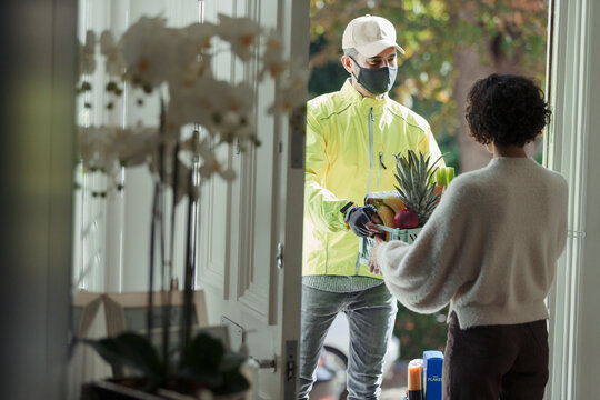 Woman Receiving Grocery Delivery From Courier In Face Mask At Door