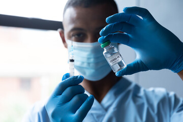 Mixed race male doctor holding covid-19 vaccination wearing face mask and gloves