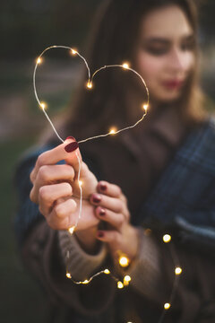 A Girl With Light LED Garland Outdoor Portrait Holds A Heart Shape. Fairy Illumination