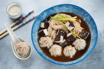Blue plate of asian miso soup with dumplings, udon noodles and tofu cheese, studio shot on a light-blue stone background