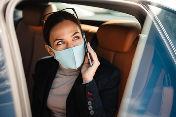 Fototapeta na wymiar Pleased businesswoman wearing face mask talking on mobile phone in car
