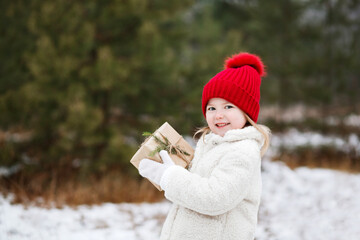 Little girl in white coat and red knitted hat smiles and holds in her hands a Christmas gift wrapped in craft paper decorated with a spruce twig outdoors in winter. Christmas and New Year concept.