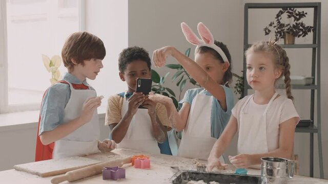 Waist up of Caucasian and Mixed-Race children playing with flour and dough, standing at kitchen table, African boy taking pictures of them with smartphone. Diverse kids in aprons having time together