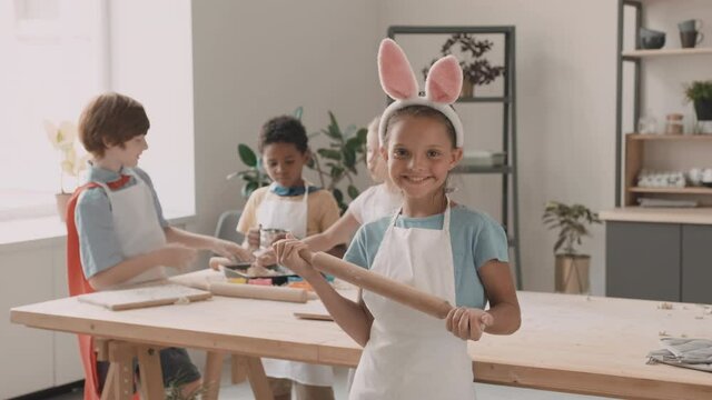 Medium long POV of Mixed-Race school girl wearing headband with bunny ears and apron, holding wooden rolling pin, standing facing diverse friends on background, turning around and smiling on camera