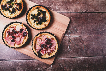 Mini individual quiche pies with filling ingredients ham,onion,spinich and cheese before egg is poured over the fillings in a baking tray on a rustic wooden background with a matt filter applied