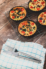 Freshly cooked mini individual quiche pies with filling ingredients ham,onion,spinich tomatoe and cheese cooling on a wire rack with knife and fork on a rustic wooden background with a matte filter 