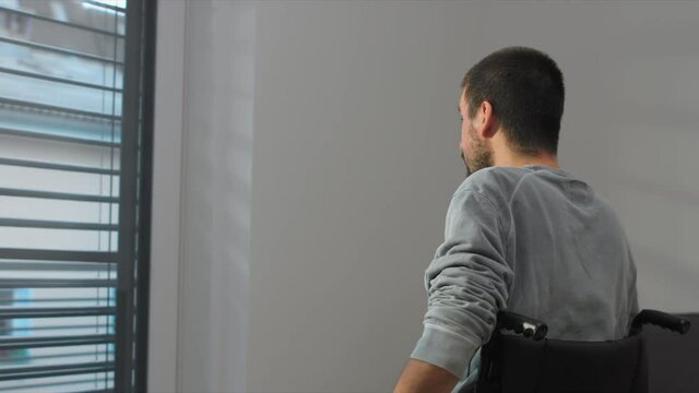 Man on wheelchair in living room looking through window. Handicapped invalid paralysed person, depressed alone at home.