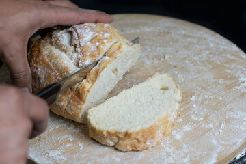 Chef's hand cutting wheat bread with a knife on floury wooden board.Rustic concept of cooking and baking on dark background.