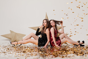 two young beautiful girls in evening dresses at a party on a light solid background with gold confetti. the concept of the celebration, the beauty of the style