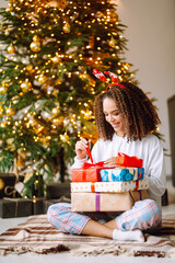 Fototapeta na wymiar Happy woman opens gifts near the Christmas tree. Young curly lady posing with presents in Christmas interior design. Fashion, celebration, holidays concept.