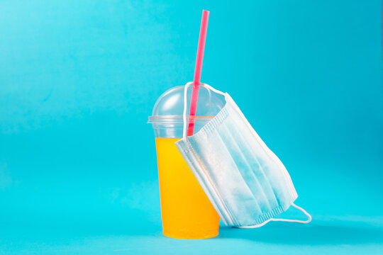 Yellow Coctail In Plastic Cup With Face Mask On Blue Background. Quarantine Catering Concept.