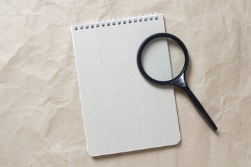 Gray notepad with white coiled spring and magnifier on a background of beige crumpled craft paper. With empty space for text and design