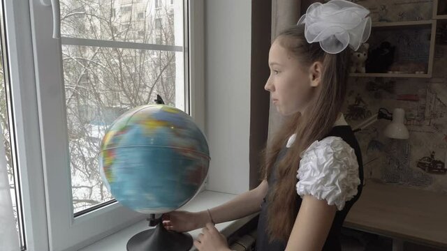 A little schoolgirl girl rotates a globe standing at the window of her room in her school uniform. She looks out the window and smiles. The concept of travel. Portrait. 4K