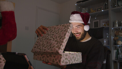 Young man smiling while opening a Christmas gift on a videocall while wearing a Christmas hat	
