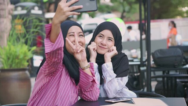 two young asian Muslim friends wear hijab taking selfies, friend meet up, sit outside coffee shop, sharing photos online post, positive friendship laughing and happiness, Ramadan holiday celebrate