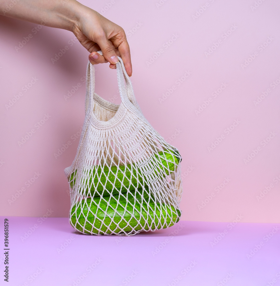 Wall mural female hand holding net bag full of fresh organic vegetables on a pink background