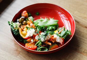 Salad with vegetables and soft Buch cheese in a red plate on a wooden table. 