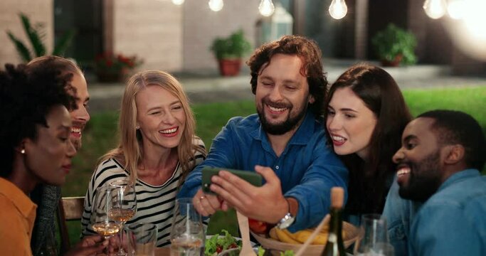Cheerful multi ethnic men and women friends sitting at table in evening and looking at photos on smartphone. Happy mixed-races happy people having fun, laughing and watching video on mobile phone.