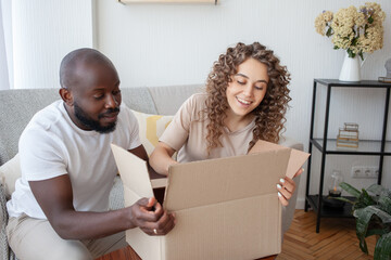 Smiling multinational couple feels happy opening a cardboard box with their long-awaited order,...