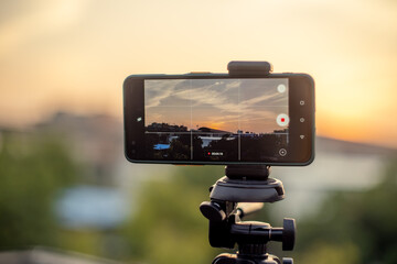 mobile phone mounted on a tripod at full extention replacing a DSLR for a professional shoot to capture a sunset with a metro station and train arriving as seen on the screen with rest out of focus