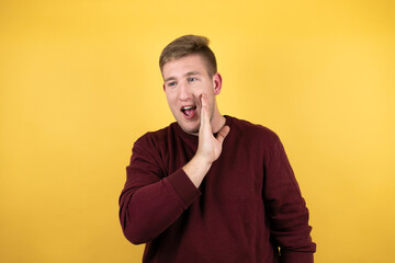 Young blonde man wearing a casual red sweater over yellow background with his hand on mouth telling secret rumor, whispering malicious talk conversation