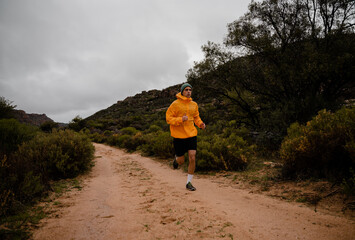 Strong young male athlete completing steep long distance run on gravel mountain path with bushes...