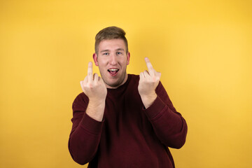 Young blonde man wearing a casual red sweater over yellow background showing middle finger doing...