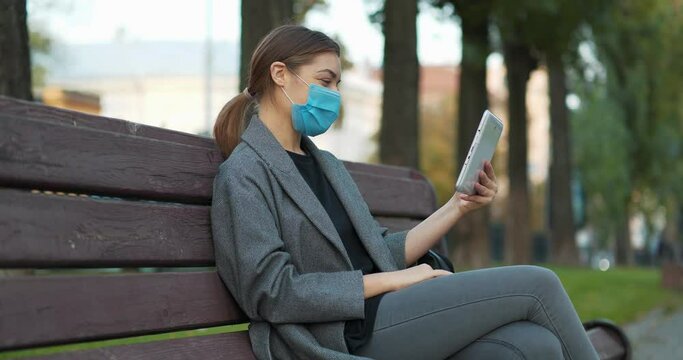 Young girl in blue medical protective mask sits on the bench in autumn city square and communicates via tablet computer. Cinema 4K 60fps panoramic video