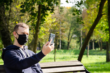 Fototapeta premium Caucasian man in protective medical mask making video call on phone and sitting on bench in the park during coronavirus outbreak with copy space. New reality. Long distance relationship concept.