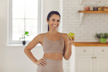 Fototapeta na wymiar Fit woman standing in the kitchen, holding fresh green apple and smiling at camera