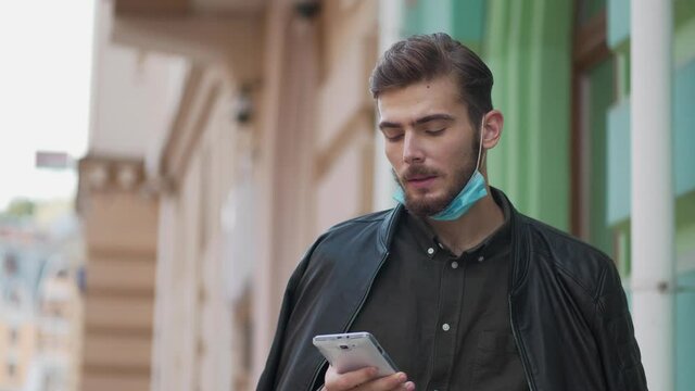 Slow Motion Young man in medical protective mask on the chin smokes a cigarette and looks in smartphone
