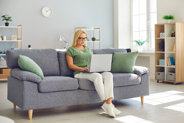 Obraz premium Young woman in eyeglasses sitting on couch and working on project using laptop computer