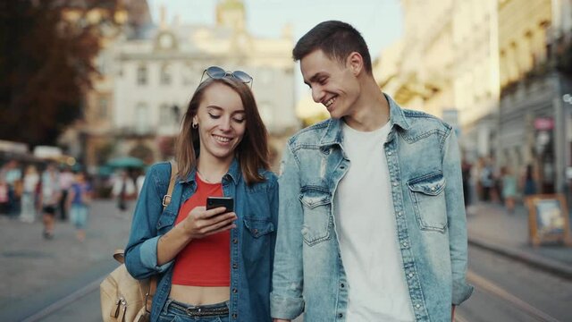 Young lovely smiling couple in casual clothes use phone walking on street old city at sunlight. Feel happy. Relationship mobile outdoors communication social media. Slow motion