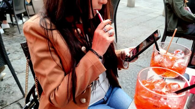 Glamour woman smoking electronic cigarette, with alcoholic drink cocktail in open bar pub. Appetizer at happy hour time with mobile phone.