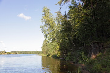 Beautiful river shore with green trees in shadow on blue sky with white cloud and calm water background at summer day, scenery Russian natural landscape view