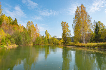 Autumn forest trees are reflected in the river water of the panoramic landscape. Blue sky with clouds.