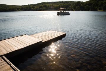 Dock over calm, shimmering sunlit lake in the summertime with boat and mountains in background