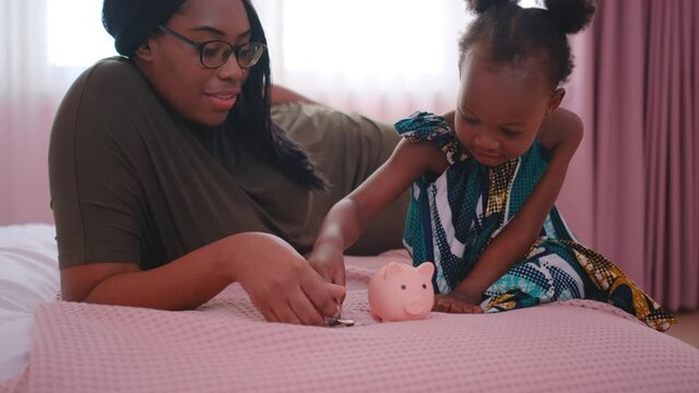 African American mother teach her daughter or little girl to collect money by put in piggy bank and stay on bed in bedroom. They look happy and enjoy together of good bonding family.