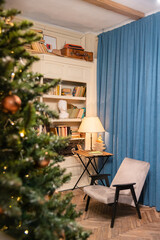 In the foreground out of focus Christmas tree decorated with Christmas balls. In the background is a place to read, a chair and a table near the bookcase. Cozy and calm atmosphere