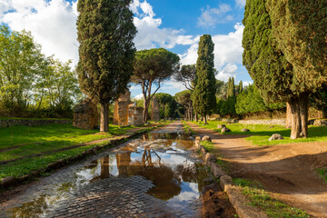 Appian Way Rome, reflections in the puddle of the remains of the tombs after the storm with the blue sky and clouds, the maritime pines and a cypress on an autumn day. Regina Viarum, Italy.