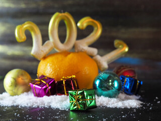 Fototapeta na wymiar small gifts in multicolored festive shiny paper lie in the snow near a ripe orange tangerine with candles in the form of 2021 numbers. horizontal image with blurred background, soft focus, free space 