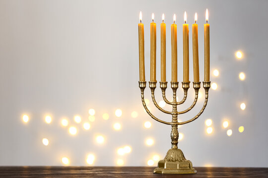 Golden menorah with burning candles on table against light grey background and blurred festive lights, space for text