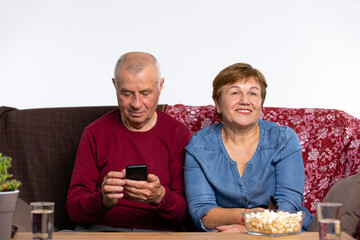 Senior spouses home on a cozy sofa. Woman is eating popcorn watching TV and man is holding a phone.