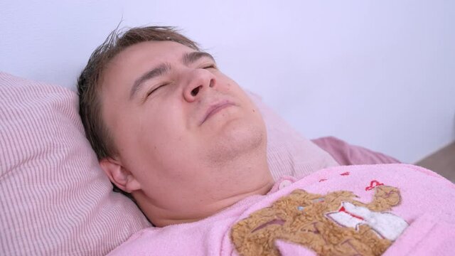 Young man in funny pink pajamas sleeps sweetly at home, mumbles in his sleep and sees exciting or pleasant dreams so he smiles and squints, close up.
