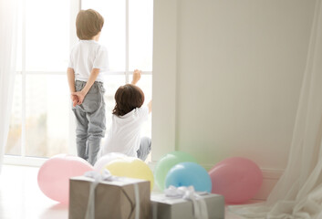 Rear view of two curious latin twin boys, children in casual wear playing at home, preparing for celebrating holiday with colorful balloons and giftboxes in the foreground