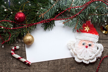 New Year and Christmas card with handmade Santa Claus, shine balls, bell and fir branches on a wooden background. A postcard with Christmas decorations and white paper.