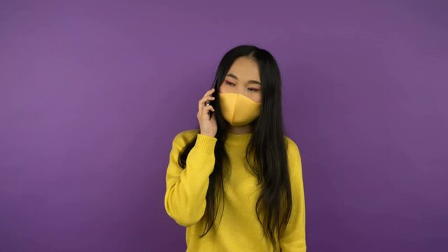 Asian woman in protective medical mask talking on the phone. lady in yellow sweater on purple isolated background. 4K