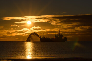 Fototapeta na wymiar Vessel engaged in dredging at sunset time. Hopper dredger working at sea. Ship excavating material from a water environment. Beautiful sunset.