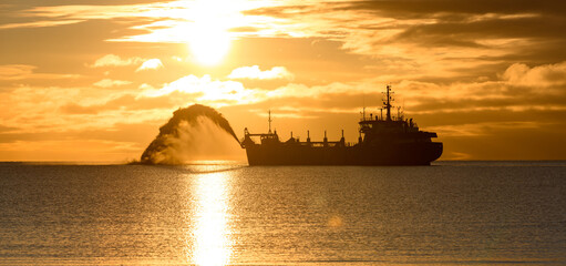 Vessel engaged in dredging at sunset time. Hopper dredger working at sea. Ship excavating material from a water environment. Beautiful sunset.